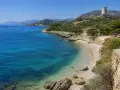 Spiaggia di Guidaloca con acque cristalline, vegetazione mediterranea e una torre antica in collina.