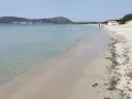 Spiaggia di Fertilia con acque cristalline, sabbia chiara e montagne sullo sfondo. Alcune persone passeggiano lungo la riva.