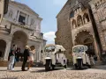 Tuk-tuk turistici pronti per esplorare cosa vedere a Bergamo in un giorno. Turisti scattano foto davanti a splendidi edifici storici.