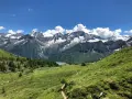 Un sentiero serpeggia tra prati verdi verso un lago alpino, con maestose montagne innevate sullo sfondo. Sembra il luogo ideale per una visita al villaggio delle marmotte.