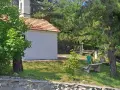 La piccola baita bianca, con tetto in tegole rosse, è circondata da alberi verdi e un campanile. Panchine di cemento invitano al riposo nel prato soleggiato.