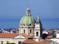 Cupola verde e campanile con orologio a Barcellona Pozzo di Gotto, con il mare azzurro sullo sfondo.
