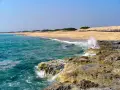 Onde cristalline del mar Ionio in Puglia si infrangono su scogli dorati, con una spiaggia sabbiosa e dune sullo sfondo.