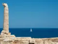 Antica colonna sulla costa degli Achei, con un veliero solitario all'orizzonte sotto un cielo azzurro.