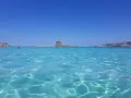 Acque cristalline e un antico torrione su un'isola: un mare bello in Italia, con gente che nuota e una barca a vela all'orizzonte.