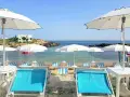 Lido Cala Pescatore: lettini azzurri e ombrelloni bianchi su una spiaggia di ciottoli, con il mare cristallino e rocce sullo sfondo.