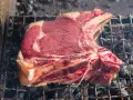 Una succulenta fiorentina di vitello, con la sua succosa carne rossa e il grasso invitante, cuoce lentamente sulla griglia ardente, sprigionando un profumo delizioso.