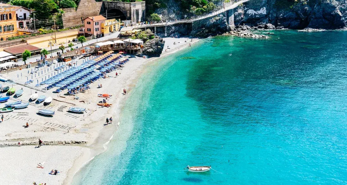 Spiaggia affollata con ombrelloni e lettini, barche sulla sabbia e mare cristallino. Un sentiero panoramico corre lungo le scogliere delle coste liguri.
