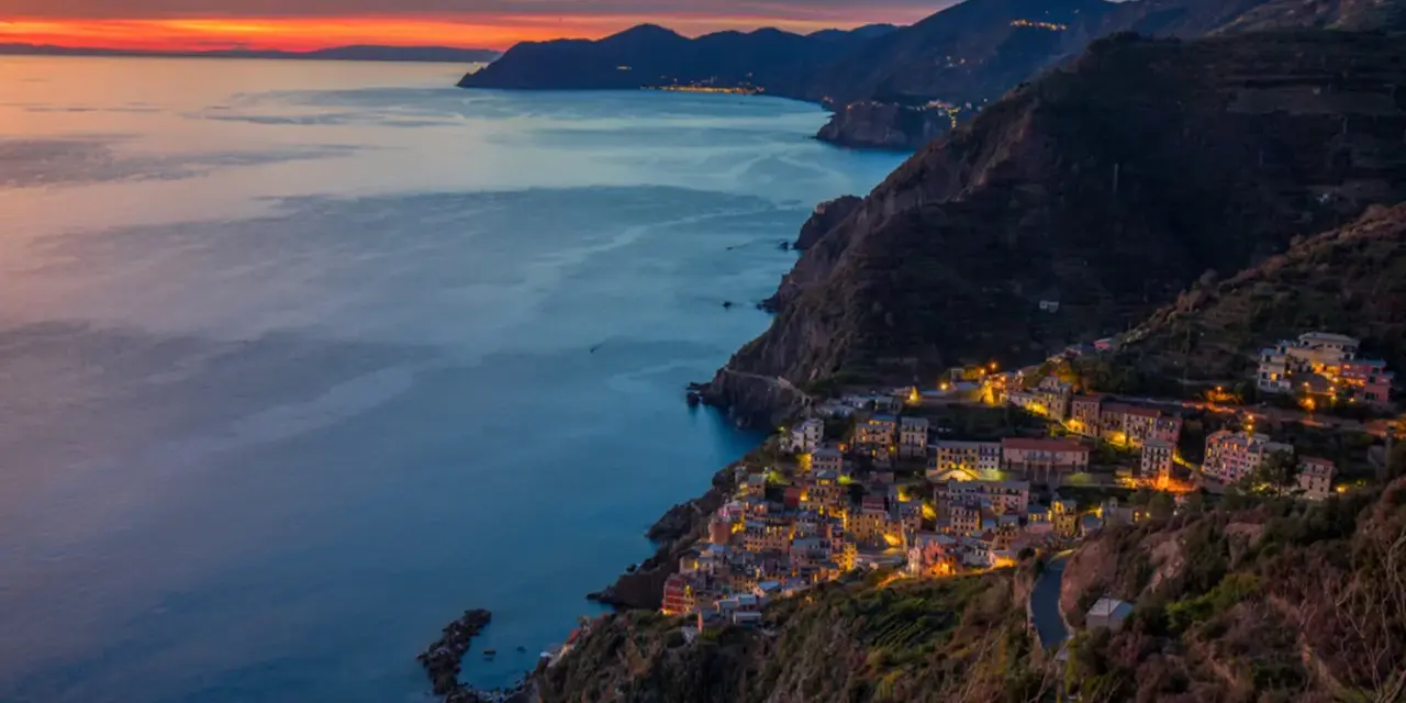 Riomaggiore cosa vedere: un villaggio colorato aggrappato alla scogliera, illuminato al tramonto, con il mare blu e le montagne sullo sfondo.