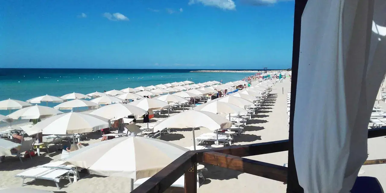Ombrelloni bianchi e lettini ordinati sulla sabbia dorata, con il mare azzurro e il cielo limpido a fare da sfondo. Un'immagine perfetta di mamire lido Marini.