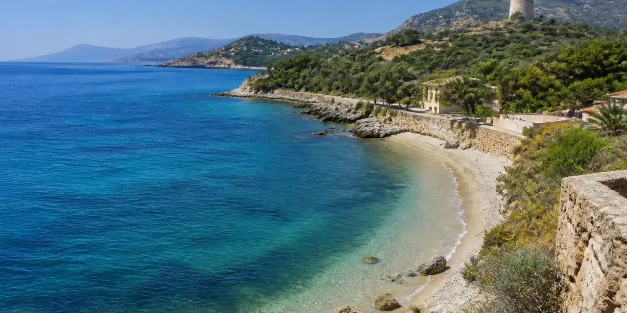 Spiaggia di Guidaloca con acque cristalline, vegetazione mediterranea e una torre antica in collina.