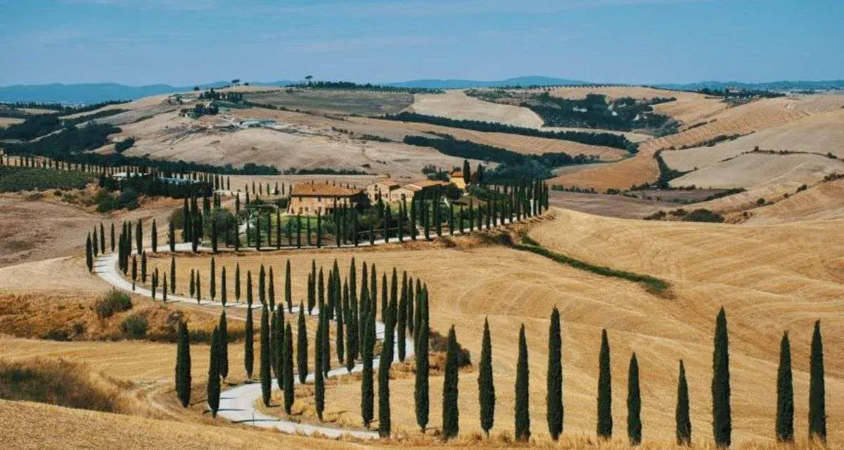 Un casale toscano circondato da cipressi lungo le strade del vino toscana, con dolci colline dorate sotto un cielo azzurro.
