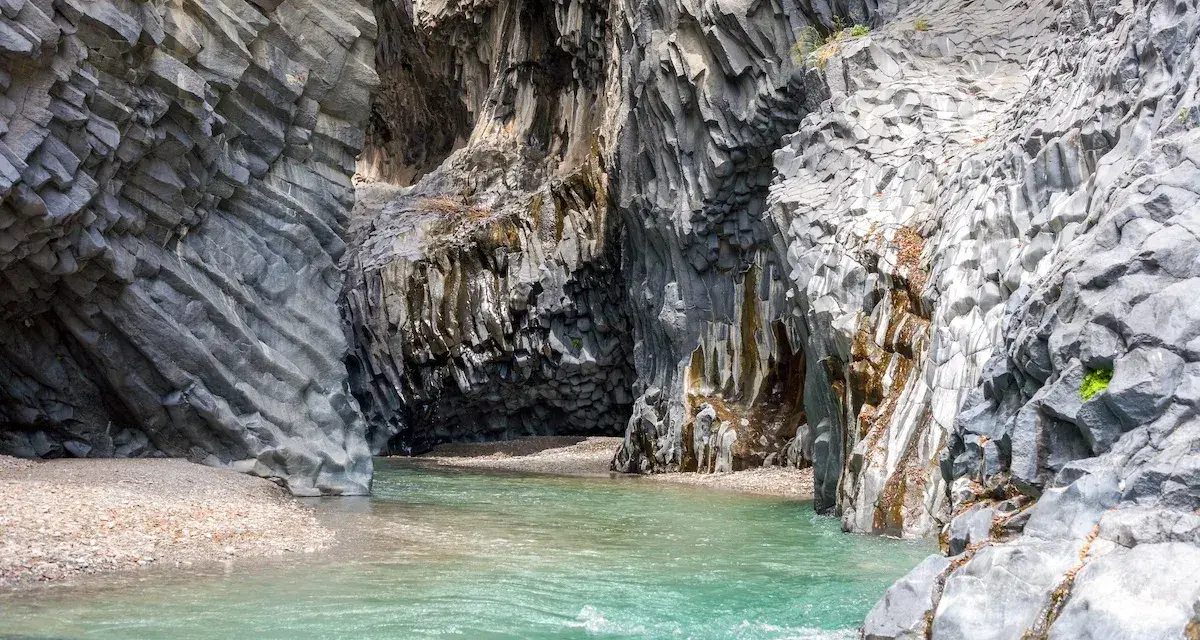 Acque cristalline scorrono tra le imponenti pareti basaltiche delle gole dell'Alcantara, creando una spiaggia naturale suggestiva.