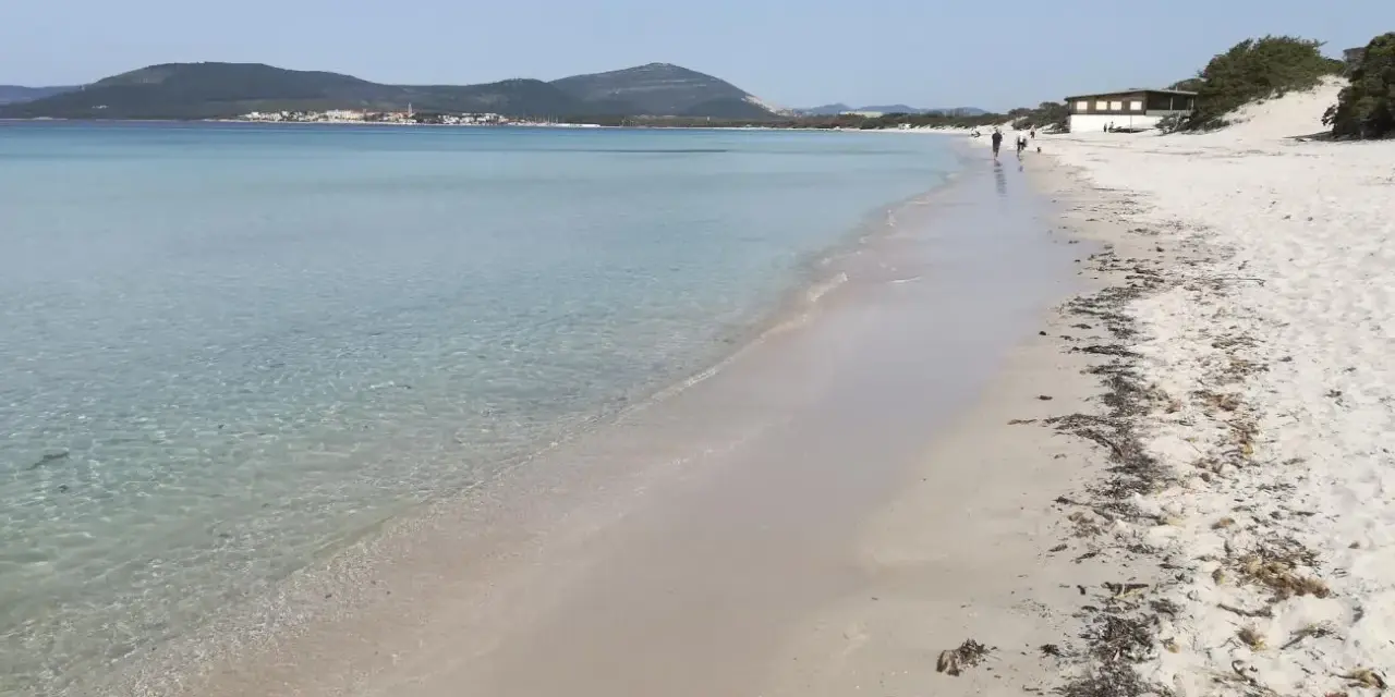 Spiaggia di Fertilia con acque cristalline, sabbia chiara e montagne sullo sfondo. Alcune persone passeggiano lungo la riva.