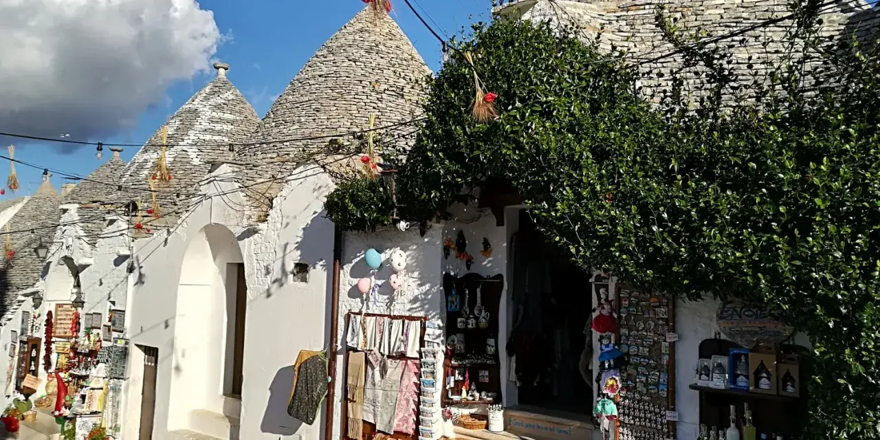 Trulli caratteristici della Valle d'Itria cosa vedere: negozi di souvenir con prodotti locali e decorazioni tipiche sotto un cielo azzurro.