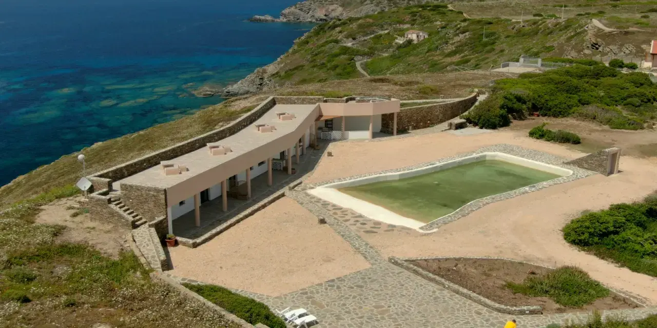 Un villaggio nurra affacciato sul mare turchese, con una piscina e lettini bianchi pronti per il relax.