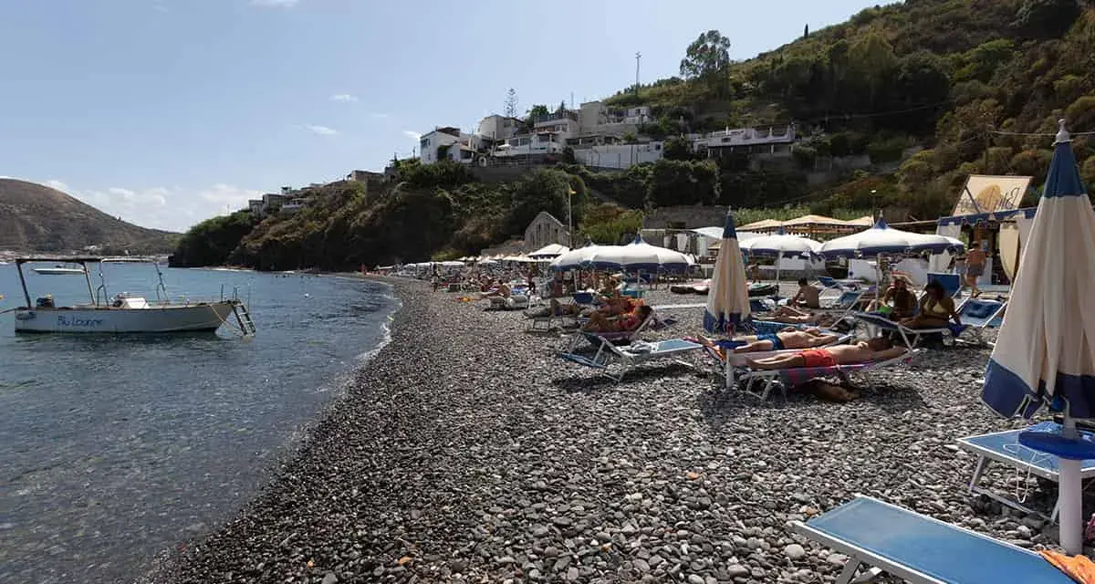 Spiaggia di ciottoli a Lipari, con lettini, ombrelloni e una barca all'ancora. La papesca beach è un luogo ideale per rilassarsi.