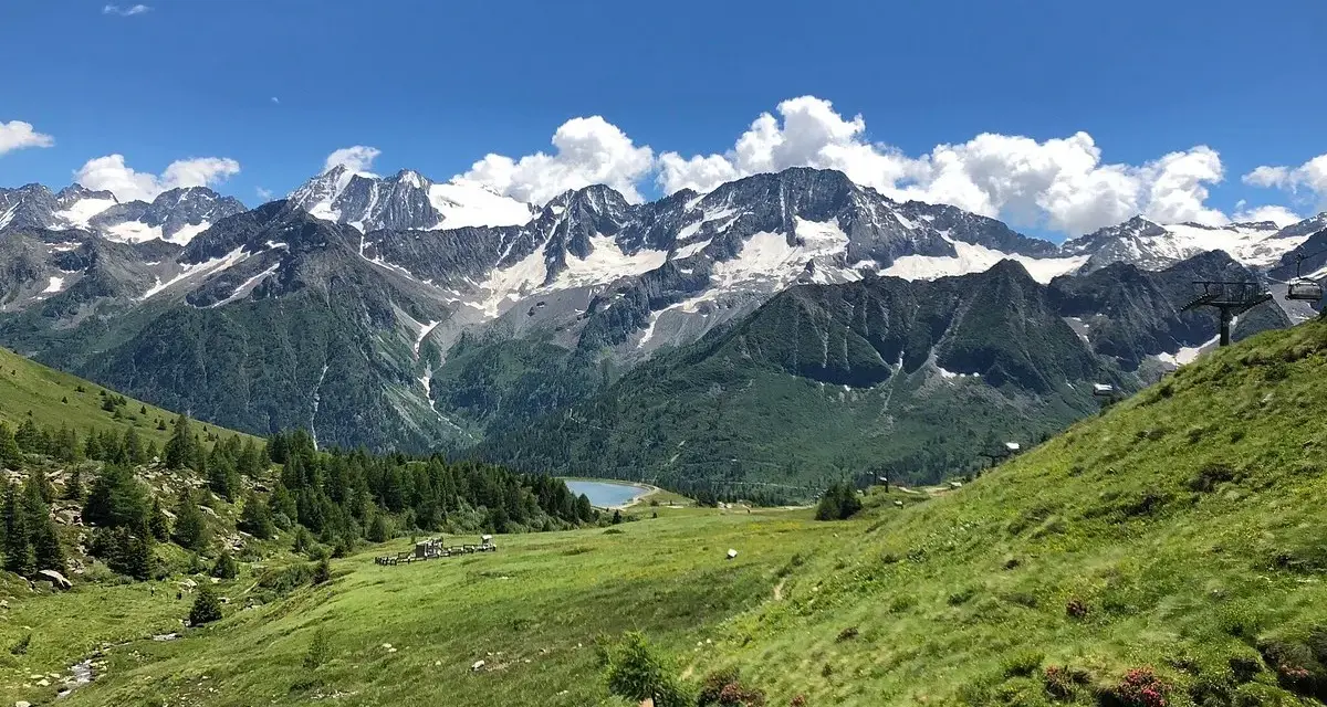 Un sentiero serpeggia tra prati verdi verso un lago alpino, con maestose montagne innevate sullo sfondo. Sembra il luogo ideale per una visita al villaggio delle marmotte.