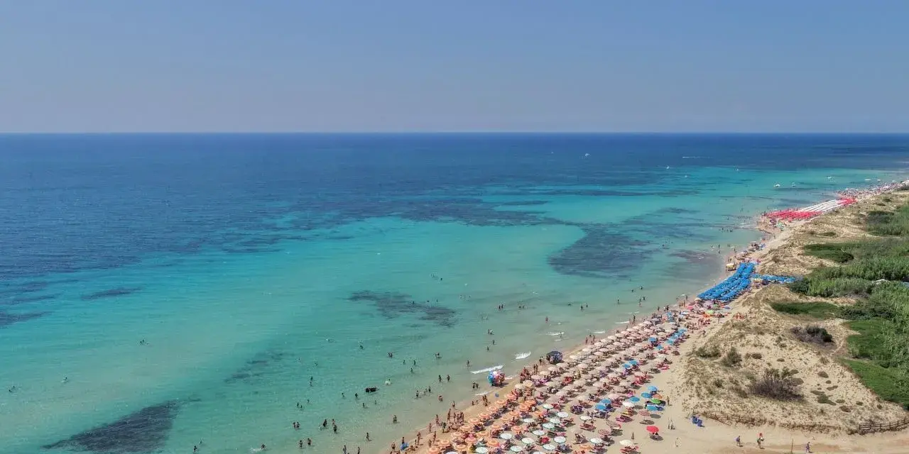 Spiaggia affollata con ombrelloni colorati e gente che nuota nelle acque cristalline di Punta Prosciutto, le Maldive del Salento.