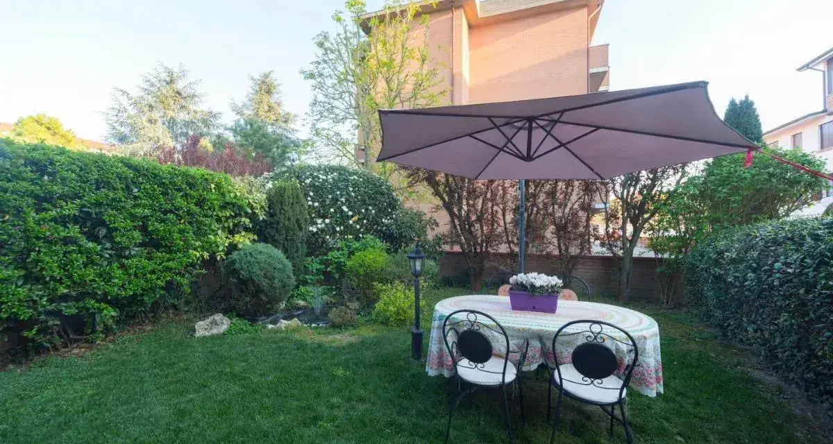 Un giardino curato con tavolo, sedie e ombrellone, circondato da siepi e alberi. Sembra l'albero dei gufi, un luogo magico per rilassarsi.