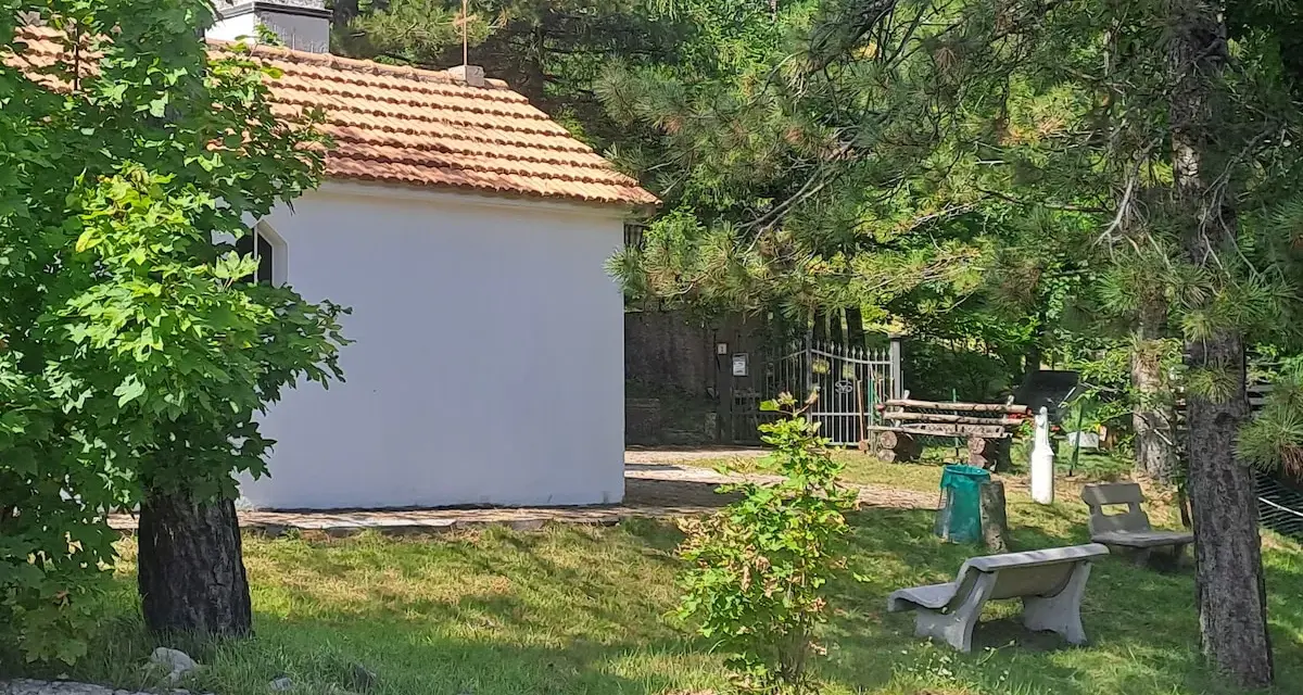 La piccola baita bianca, con tetto in tegole rosse, è circondata da alberi verdi e un campanile. Panchine di cemento invitano al riposo nel prato soleggiato.