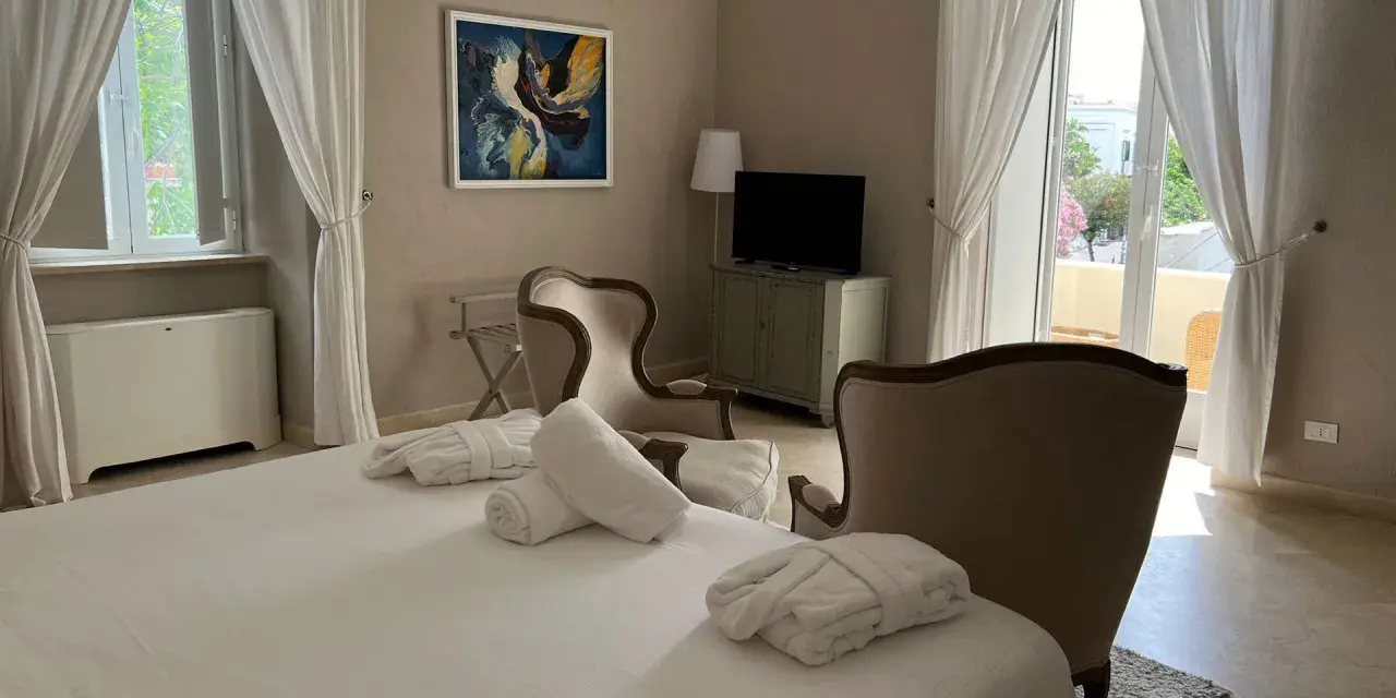 Camera accogliente al relais Corte degli Aragonesi, con letto, accappatoi, poltrone e vista su un balcone fiorito.
