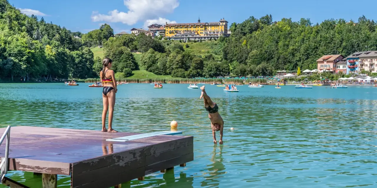 Ragazzo si tuffa in un lago balneabile, mentre una ragazza osserva dal pontile. Altri bagnanti e pedalò popolano le acque.