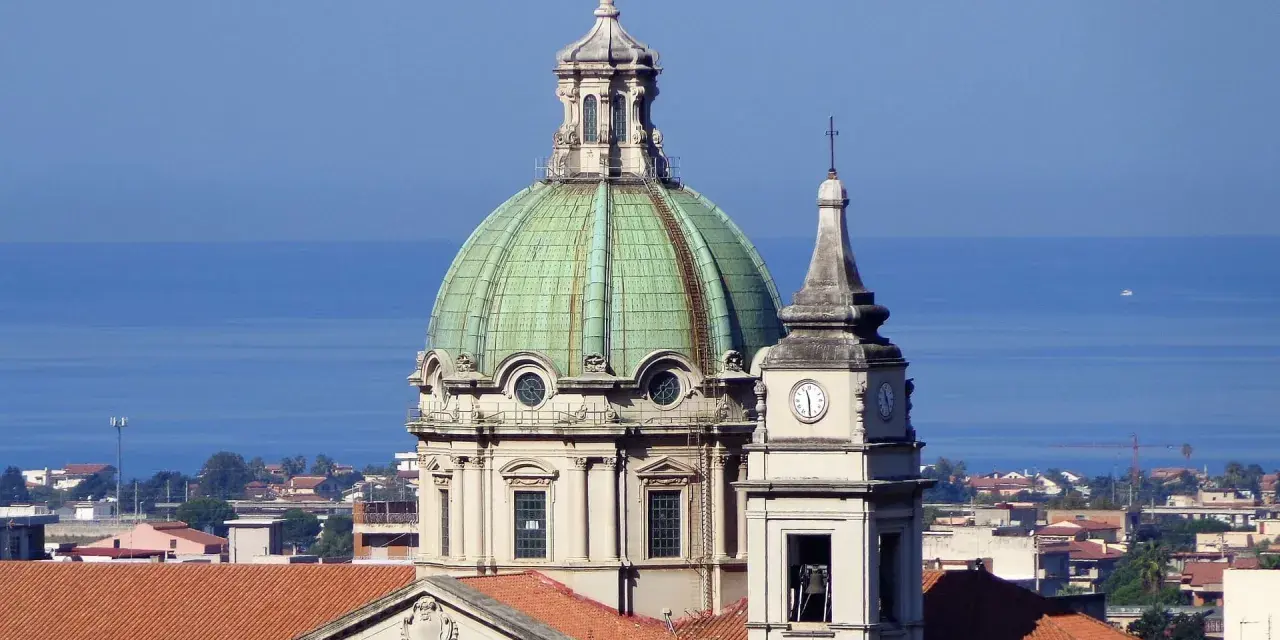 Cupola verde e campanile con orologio a Barcellona Pozzo di Gotto, con il mare azzurro sullo sfondo.