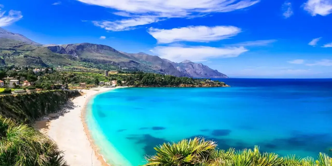 Una baia incantevole con acque cristalline e sabbia dorata, tra le spiagge più belle in Sicilia. Montagne verdi sullo sfondo.