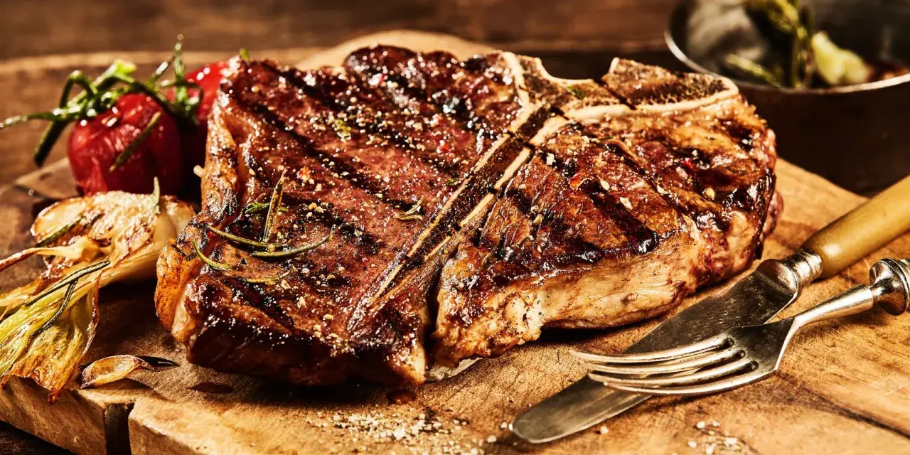 Una succulenta fiorentina, cotta alla perfezione, riposa su un tagliere di legno, pronta per essere gustata. Accanto, pomodorini e aglio arrostiti.