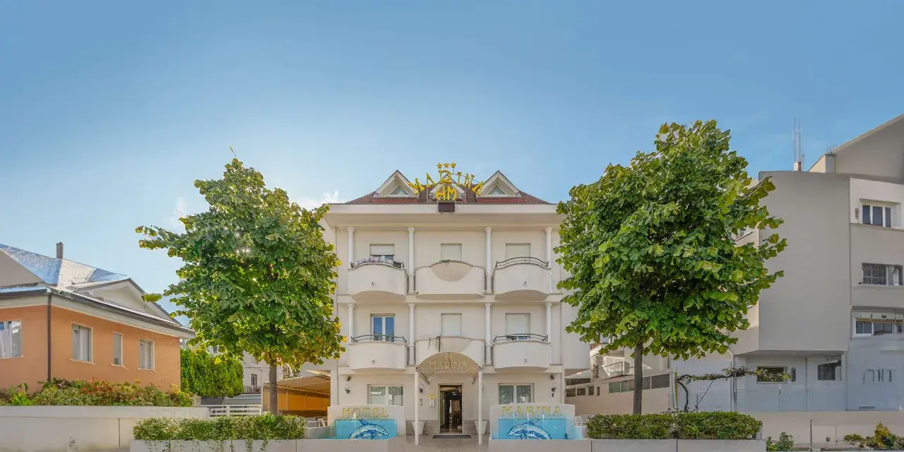 Hotel Marina Riccione: facciata bianca con insegna dorata, alberi verdi e fontane. Le recensioni lo descrivono come un'ottima scelta.