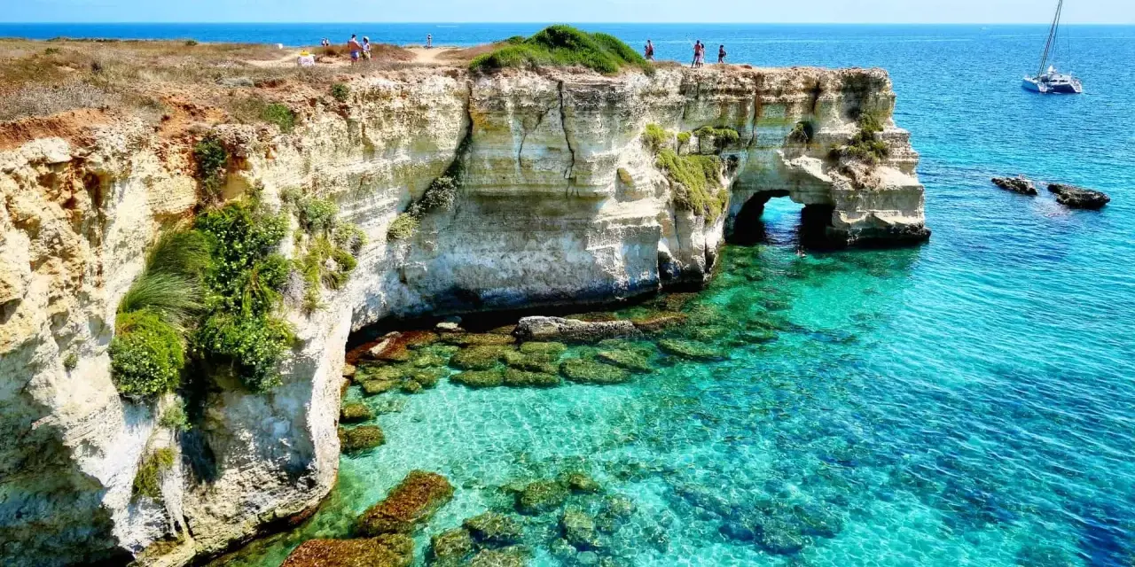 Scogliere bianche e mare cristallino sul litorale adriatico. Turisti ammirano il panorama, un catamarano naviga in lontananza.