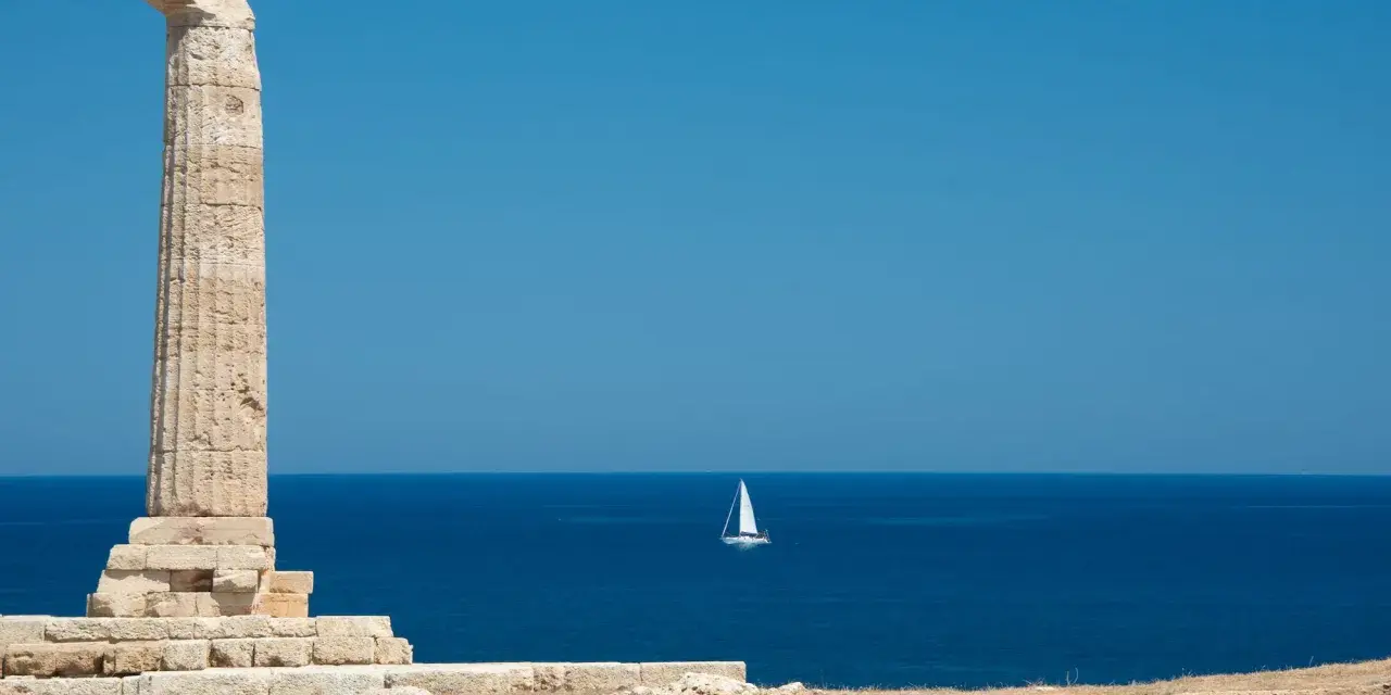 Antica colonna sulla costa degli Achei, con un veliero solitario all'orizzonte sotto un cielo azzurro.
