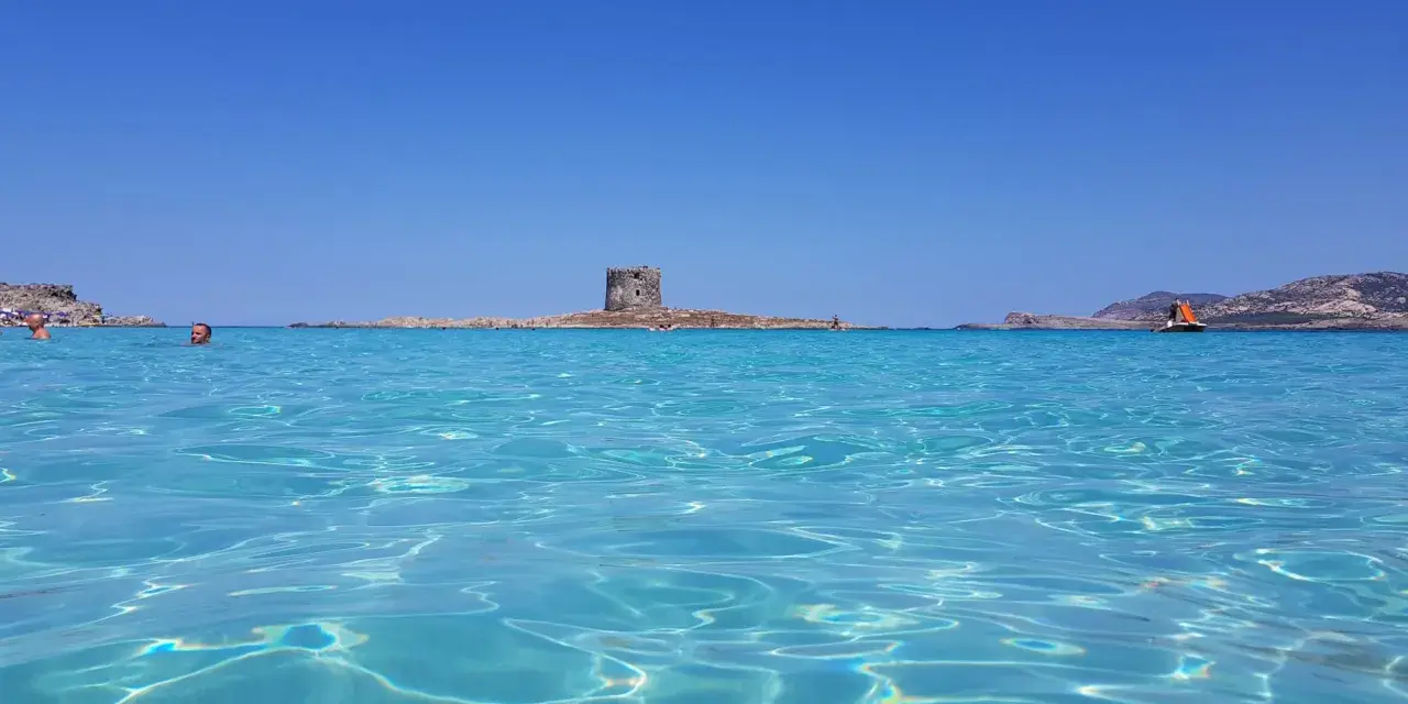 Acque cristalline e un antico torrione su un'isola: un mare bello in Italia, con gente che nuota e una barca a vela all'orizzonte.