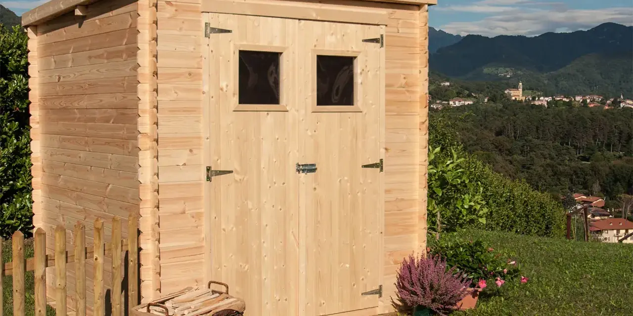 La casetta in campagna, in legno chiaro, con due finestrelle e un cesto di legna, si affaccia su un panorama collinare con un borgo in lontananza.