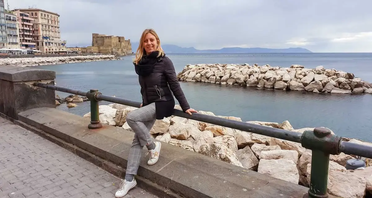 Donna sorridente sul lungomare di Napoli, con Castel dell'Ovo sullo sfondo. Perfetto per scoprire cosa vedere a Napoli in 3 giorni.