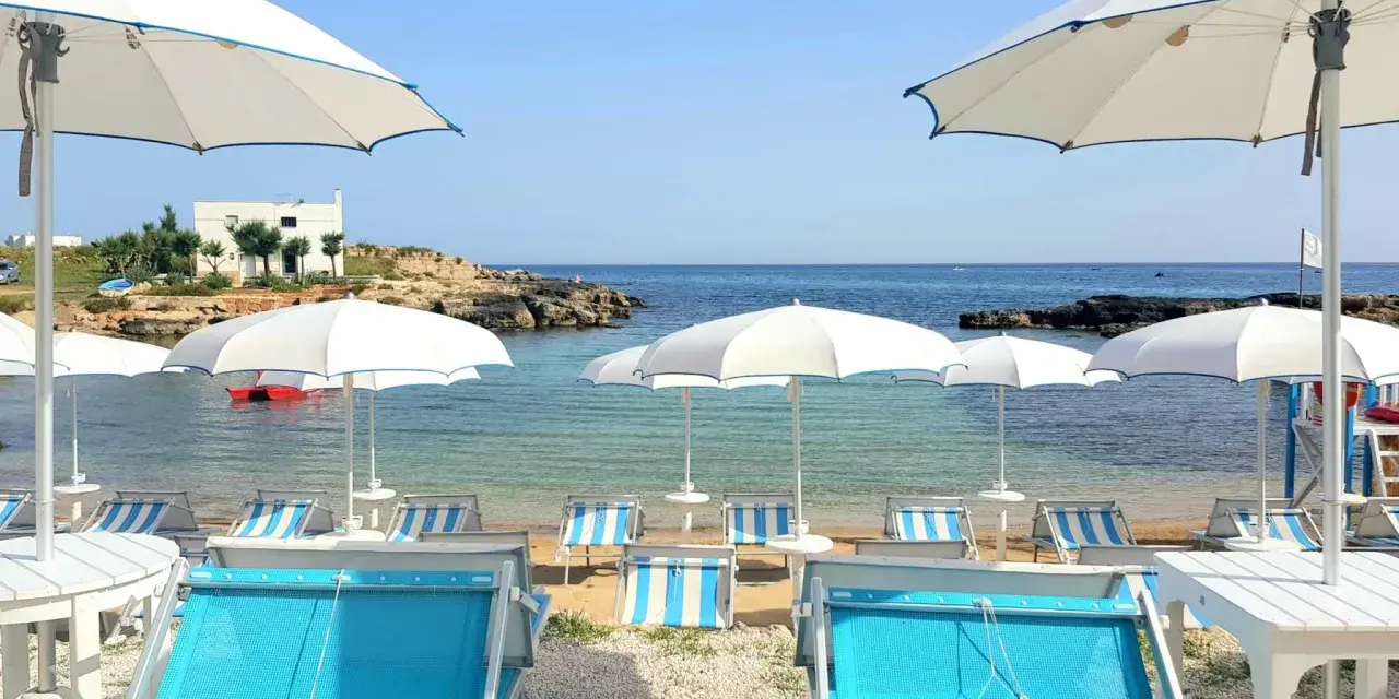 Lido Cala Pescatore: lettini azzurri e ombrelloni bianchi su una spiaggia di ciottoli, con il mare cristallino e rocce sullo sfondo.