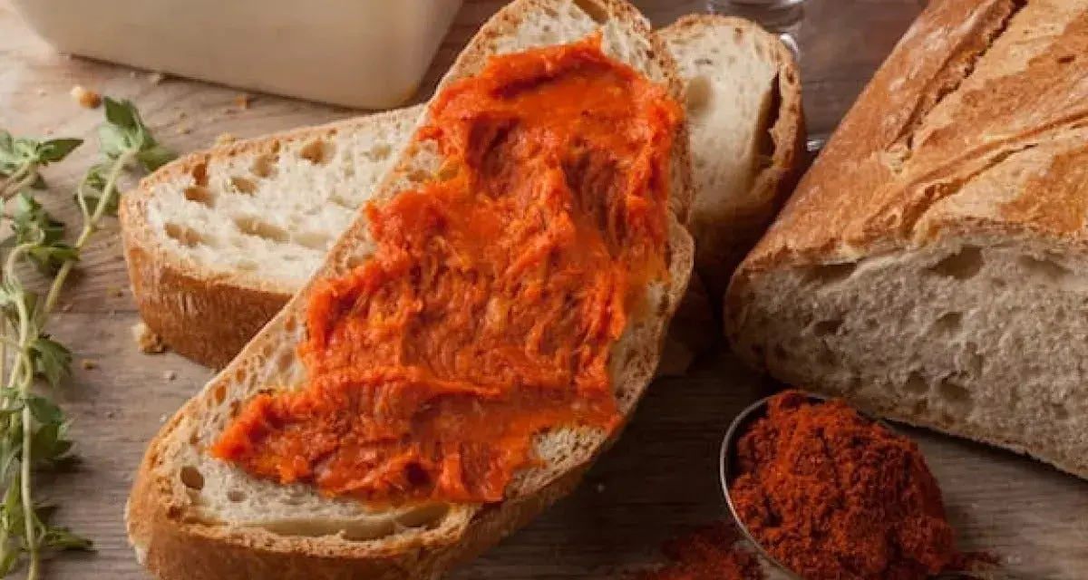 Fetta di pane con 'nduja, uno dei piatti tipici della Calabria, accanto a una ciotola con la crema e un cucchiaino di peperoncino in polvere.