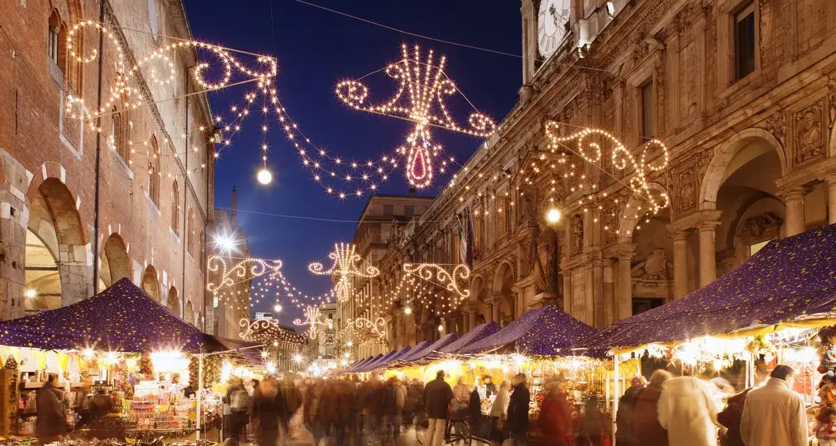 Una strada illuminata da lucine natalizie, con bancarelle e gente che passeggia: ecco i mercatini di Natale più belli.