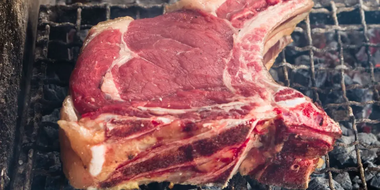 Una succulenta fiorentina di vitello, con la sua succosa carne rossa e il grasso invitante, cuoce lentamente sulla griglia ardente, sprigionando un profumo delizioso.