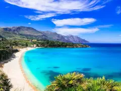 Una baia incantevole con acque cristalline e sabbia dorata, tra le spiagge più belle in Sicilia. Montagne verdi sullo sfondo.
