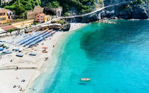 Spiaggia affollata con ombrelloni e lettini, barche sulla sabbia e mare cristallino. Un sentiero panoramico corre lungo le scogliere delle coste liguri.