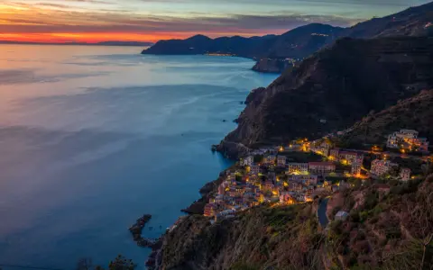 Riomaggiore cosa vedere: un villaggio colorato aggrappato alla scogliera, illuminato al tramonto, con il mare blu e le montagne sullo sfondo.