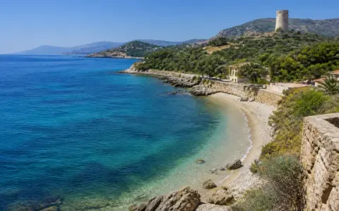 Spiaggia di Guidaloca con acque cristalline, vegetazione mediterranea e una torre antica in collina.