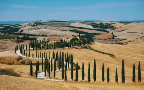 Un casale toscano circondato da cipressi lungo le strade del vino toscana, con dolci colline dorate sotto un cielo azzurro.