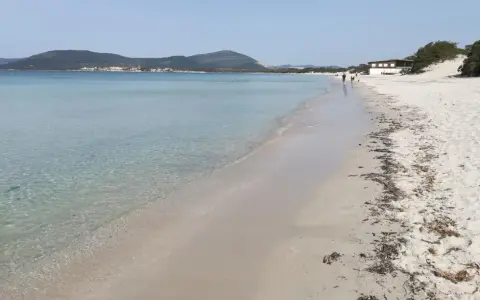 Spiaggia di Fertilia con acque cristalline, sabbia chiara e montagne sullo sfondo. Alcune persone passeggiano lungo la riva.