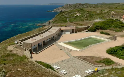 Un villaggio nurra affacciato sul mare turchese, con una piscina e lettini bianchi pronti per il relax.