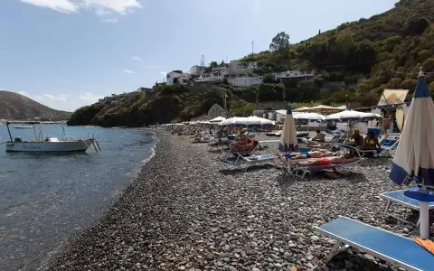 Spiaggia di ciottoli a Lipari, con lettini, ombrelloni e una barca all'ancora. La papesca beach è un luogo ideale per rilassarsi.