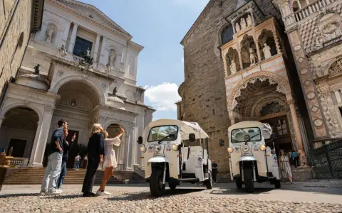 Tuk-tuk turistici pronti per esplorare cosa vedere a Bergamo in un giorno. Turisti scattano foto davanti a splendidi edifici storici.