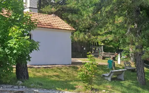 La piccola baita bianca, con tetto in tegole rosse, è circondata da alberi verdi e un campanile. Panchine di cemento invitano al riposo nel prato soleggiato.
